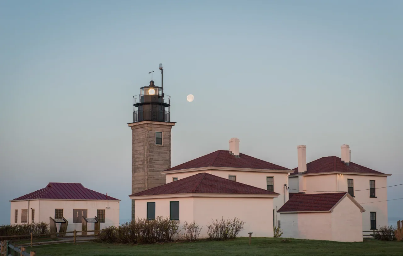 Beavertail Lighthouse and its red-roofed buildings at dawn, with the moon beside the lit lantern.