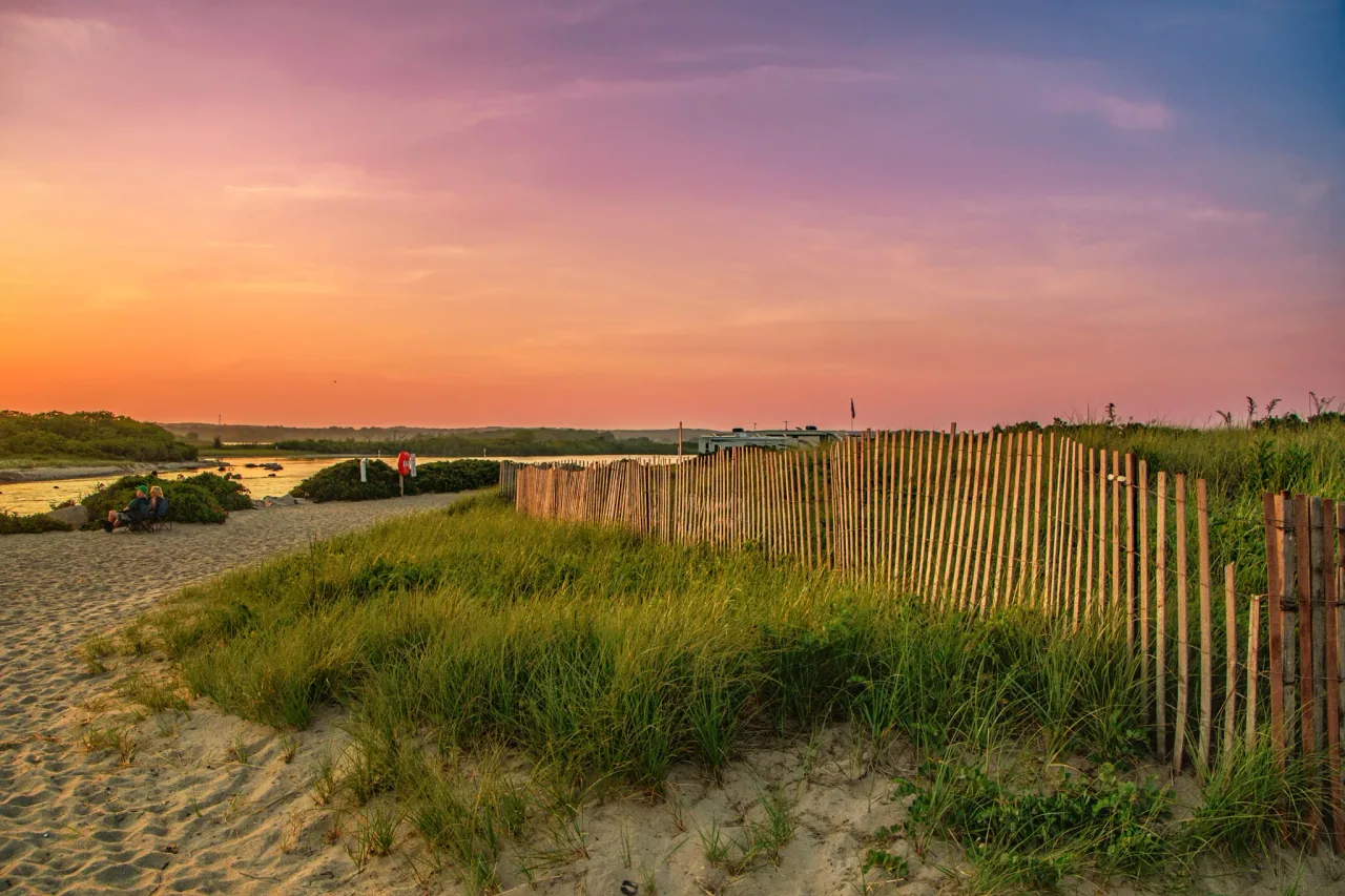 An evening sky glows above the sandy path, dune fence, and channel at Charlestown Breachway.