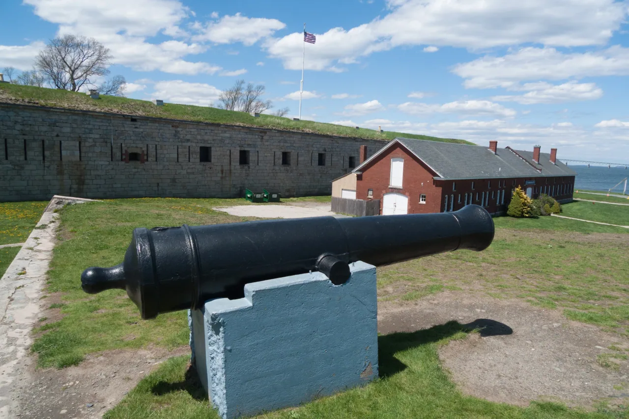 A cannon stands before the stone walls and buildings of Fort Adams under a blue sky.