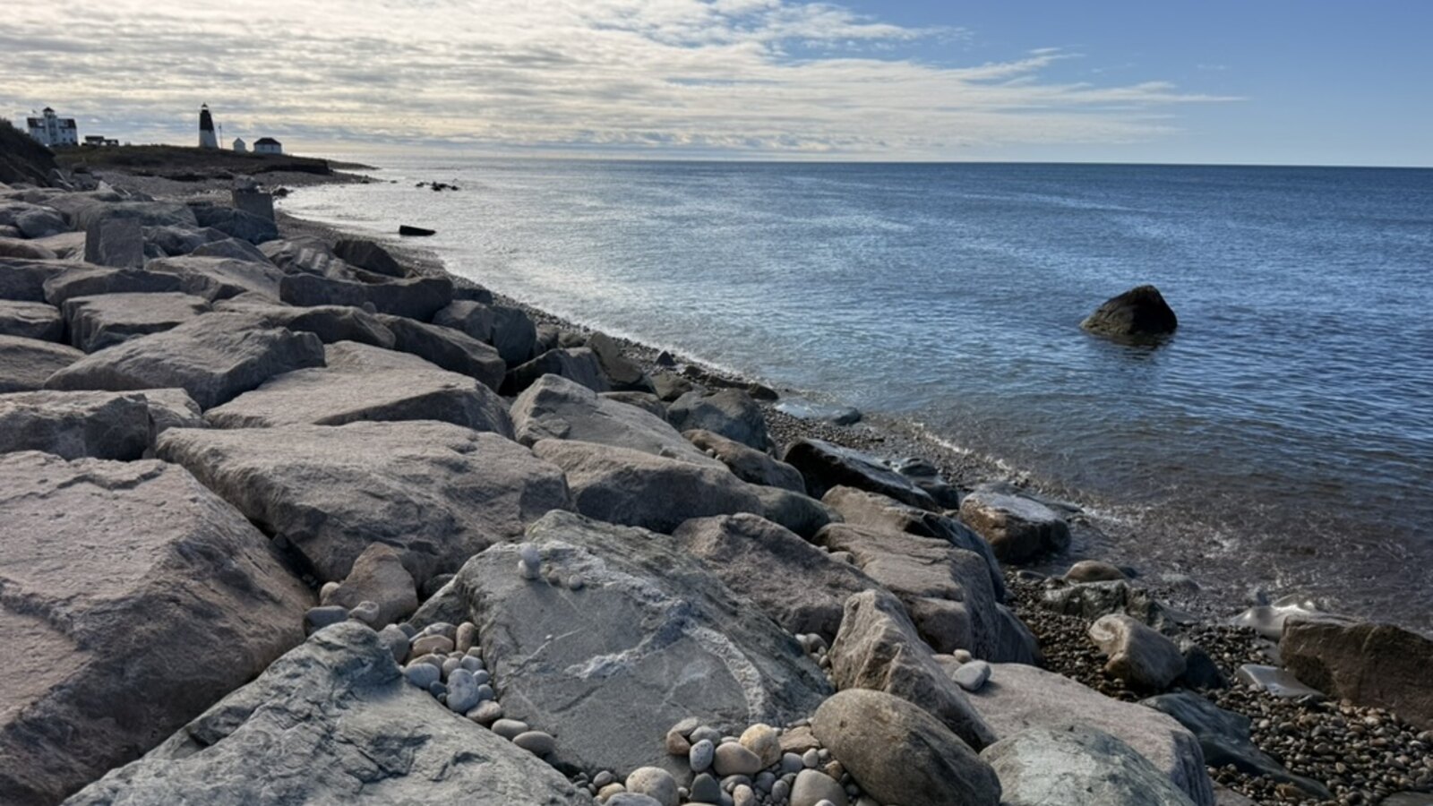 A rocky Rhode Island shoreline looking toward the ocean with a lighthouse and coastal buildings in the distance.